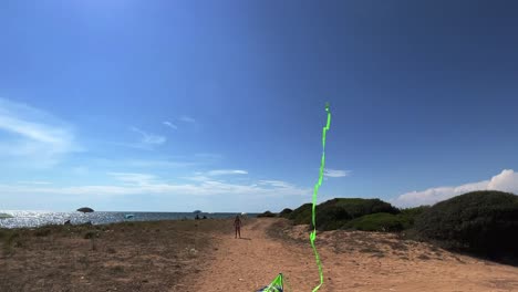 niña en vacaciones de verano jugando con cometa de juguete verde con cola larga en el cielo azul sosteniendo manijas y sentada en una silla pequeña en la playa de arena