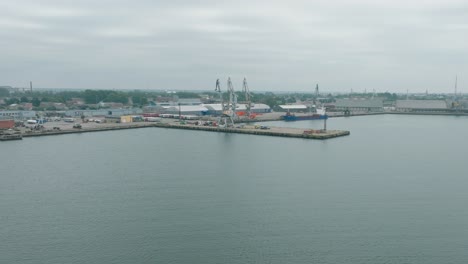 aerial establishing view of port cranes and empty loading docks at port of liepaja , liepaja city in the background, overcast summer day, wide orbiting drone shot moving right