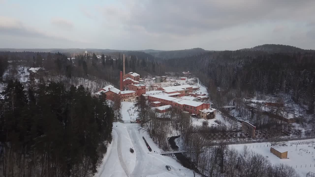 acercándose a una gran fábrica y fábrica de papel de ladrillo rojo rodeada de bosques y colinas invernales y dos torres de chimenea altas