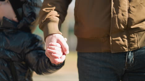 a close up of an adult and a child walking hand in hand, the child raises his ight hand up, the adult is dressed in a brown jacket and jeans, while the child is wearing a shiny black jacket