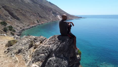 barefoot meditating man sitting on the edge of a cliff contemplating the panoramic view of the amphitheater bay of loutro