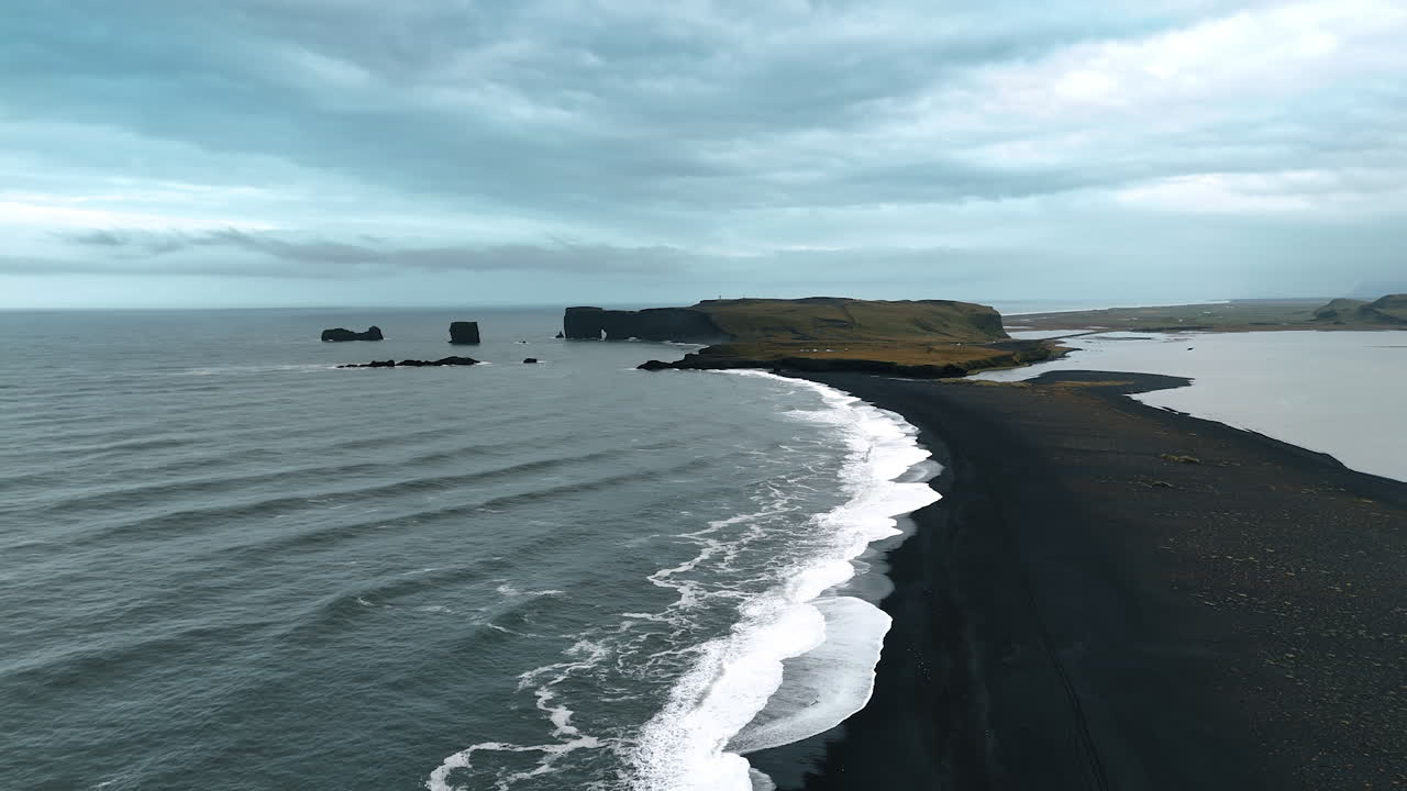 Thin line of the beach with black sand among the waters of the ocean. Waterfront view with the rock at the back. Aerial perspective on the Iceland scenery.