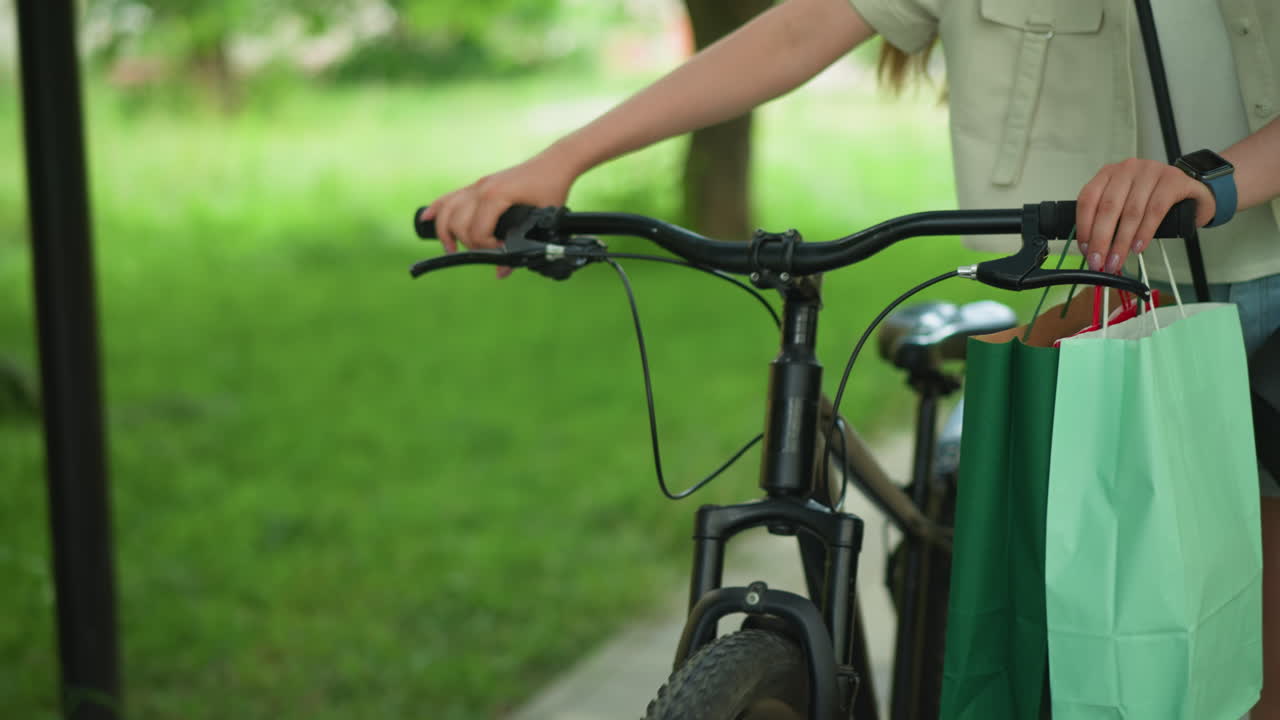 mujer paseando con la mano en un manillar de bicicleta, bolsas de compras verdes y menta colgando del mango, ajusta su bolsa negra de cuerpo cruzado, con un fondo borroso de hierba verde