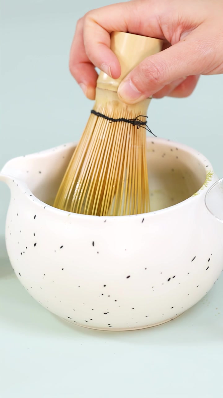 Primo piano di una mano che sbatte il tè verde matcha in una ciotola di ceramica maculata sotto una luce soffusa