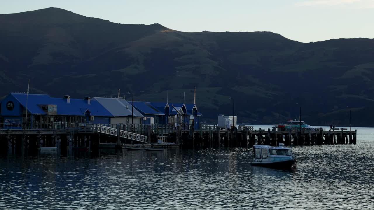 A boat moves towards a pier against a backdrop of mountains during sunset, creating a serene and tranquil scene