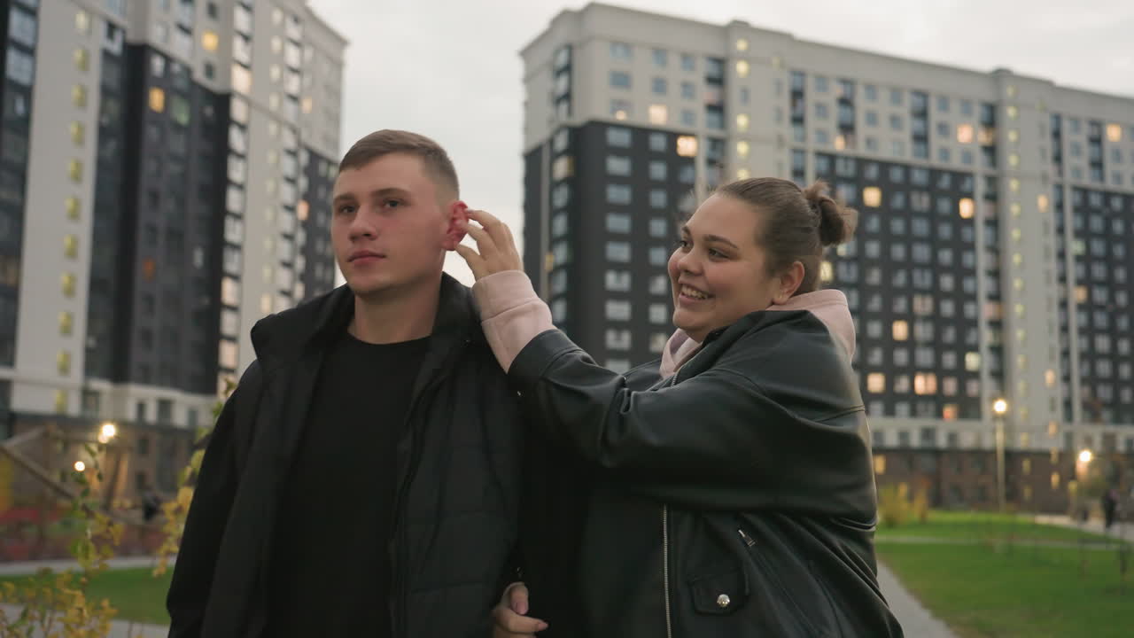 Friends walking side by side as cheerful lady playfully tickles man trying not to laugh until he gives in with big smile while evening lights glow behind tall residential buildings and green park