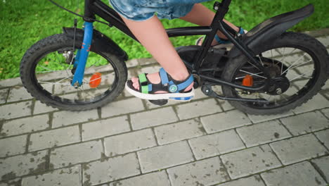 a close-up of a child's leg on a bicycle, riding in an uneven pattern, with a woman's foot seen following behind