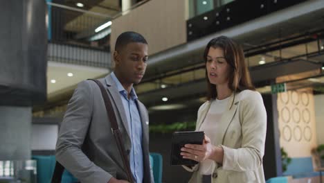 Diverse-businessman-and-businesswoman-talking-and-using-tablet-in-lobby-of-modern-office