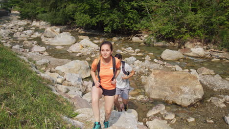 Two-tourists-hiking-along-the-rocky-mountain-stream-bed,-handheld-shot