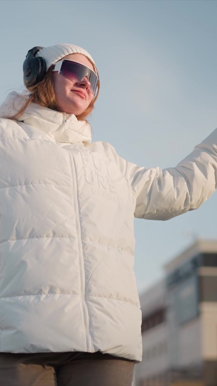 Young woman wearing white puffer jacket with FUTURE text black headphones and sunglasses moves her arms against outdoor urban backdrop. Her body language suggests casual movement or dance