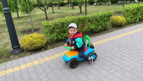 cheerful caucasian toddler in jacket and cap riding toy car outdoors. happy active kid on the walk in the park.