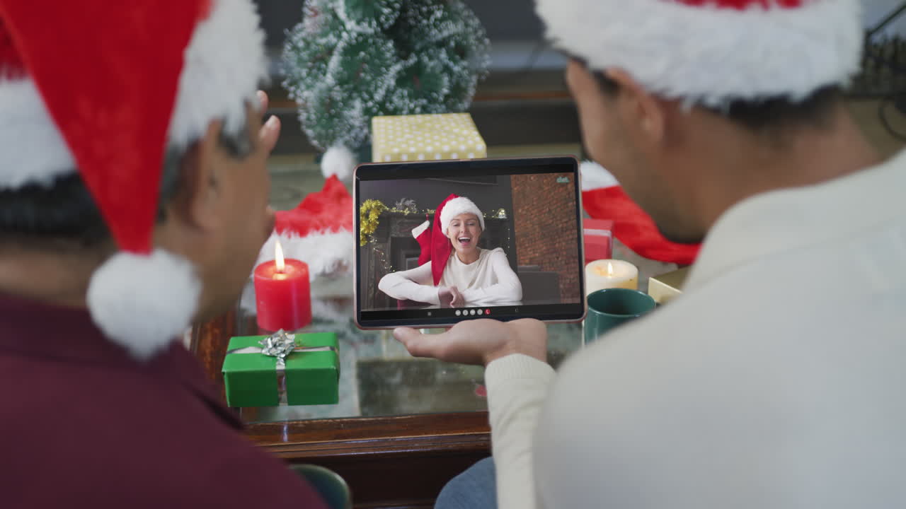 padre y hijo biraciales agitando y usando una tableta para una videollamada de navidad con una mujer en la pantalla
