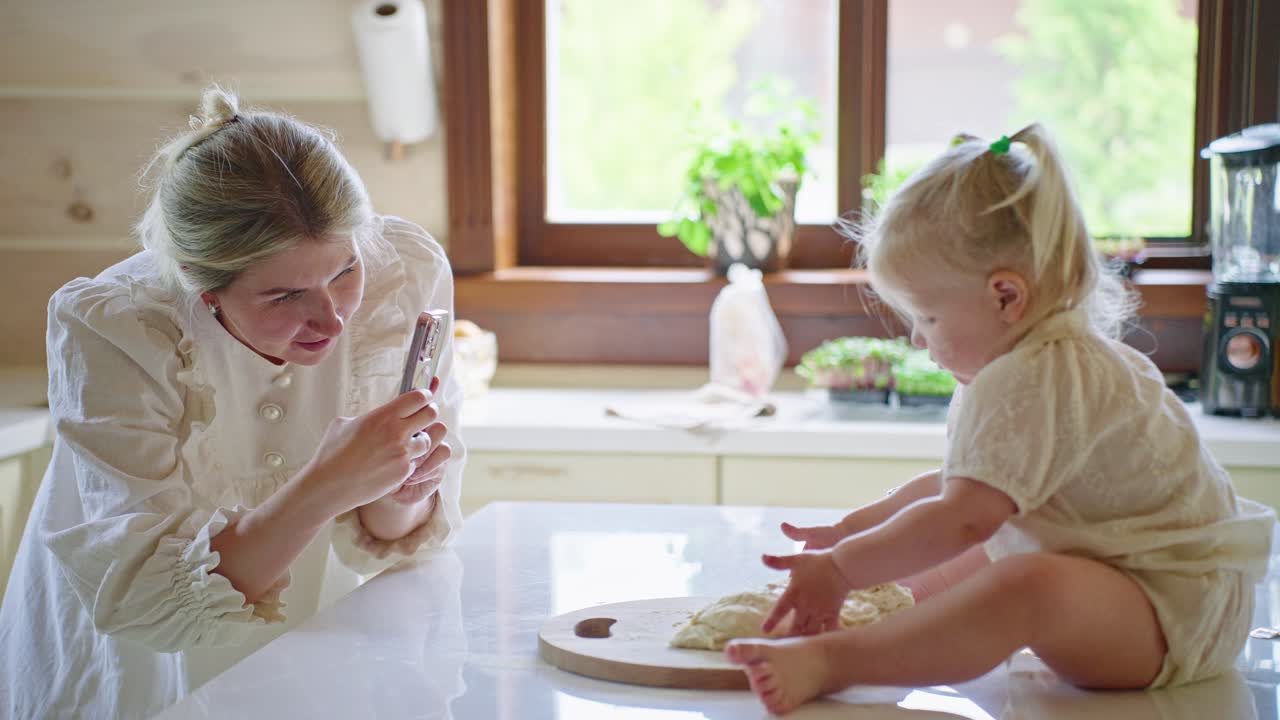 Mother photographs daughter playing with dough in kitchen
