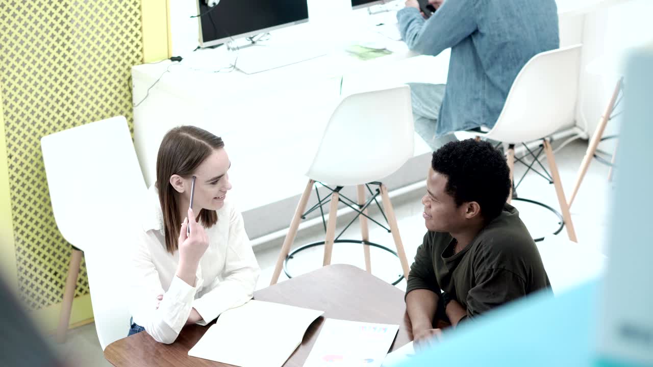 a través de la estantería vista de una bonita chica de la universidad sonriendo mientras explica la tarea a su compañera de clase africana