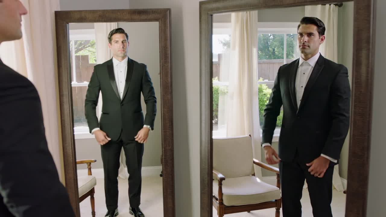 Un uomo in smoking si controlla allo specchio, abbottonando la giacca e sistemandosi il papillon prima dell'evento.