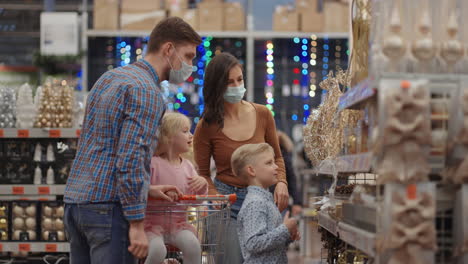 four people mom dad and two kids with a shopping cart. a happy family in medical masks in the store buys christmas decorations and gifts in slow motion