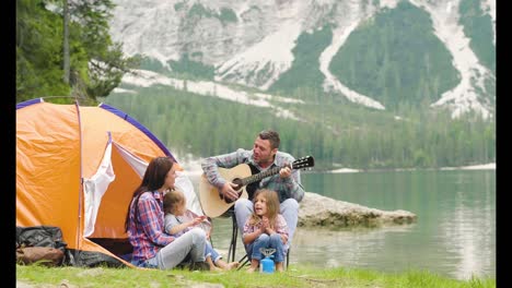 a happy family camping at the lake, playing the guitar and singing a song together in front of the tent.