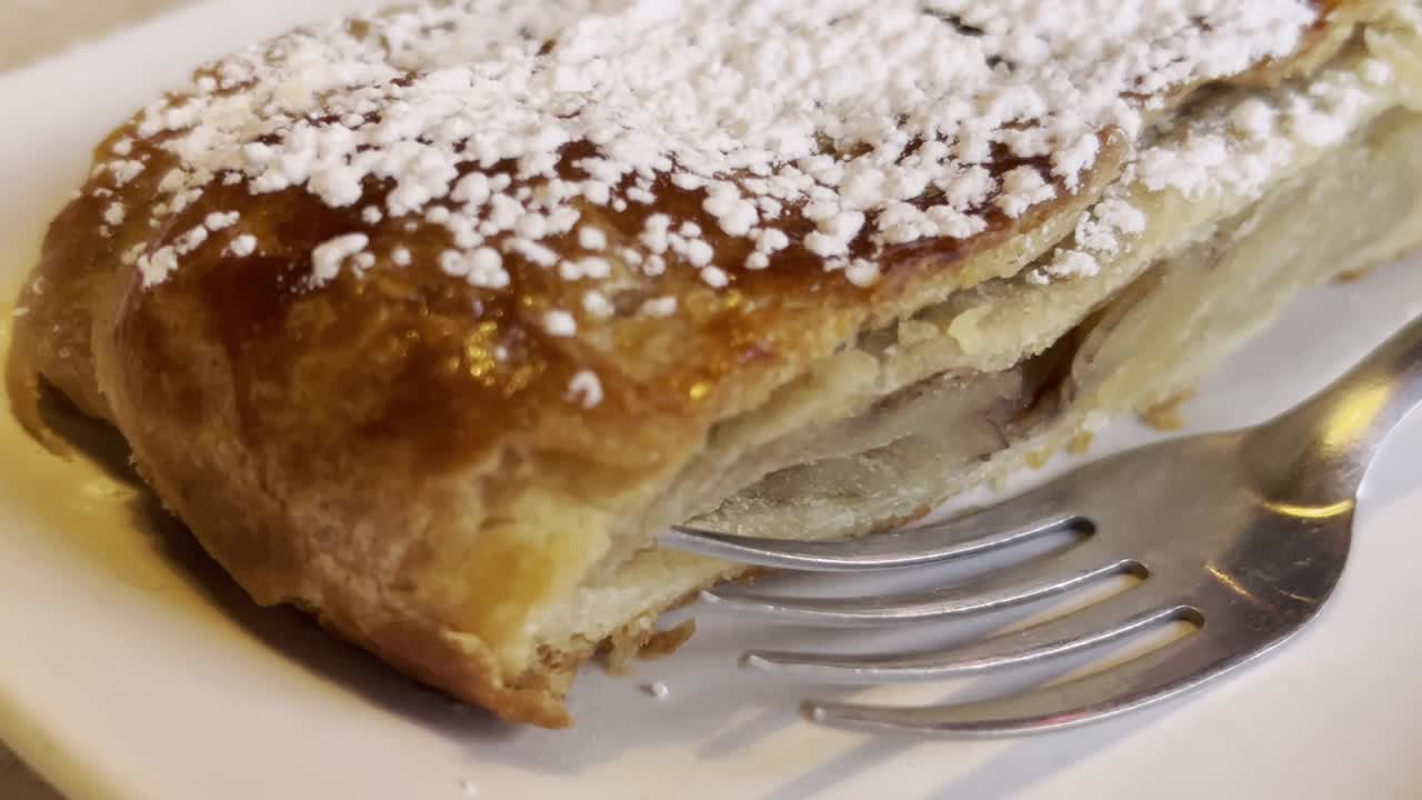 Puff pastry filled with sweet apple, Strudel recipe, dusted with icing sugar, resting on a white plate. Enjoying a delicious dessert in a cozy restaurant setting in Mexico, slow reveal