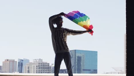 hombre afroamericano con gafas de sol sosteniendo la bandera de la lbgt mientras está de pie cerca de la ventana