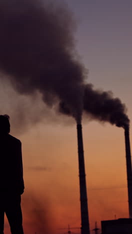 massive dark smoke going from pipes at sunset. silhouette of a man standing against the evening sky with dirty dense emissions in the air. air pollution. vertical video