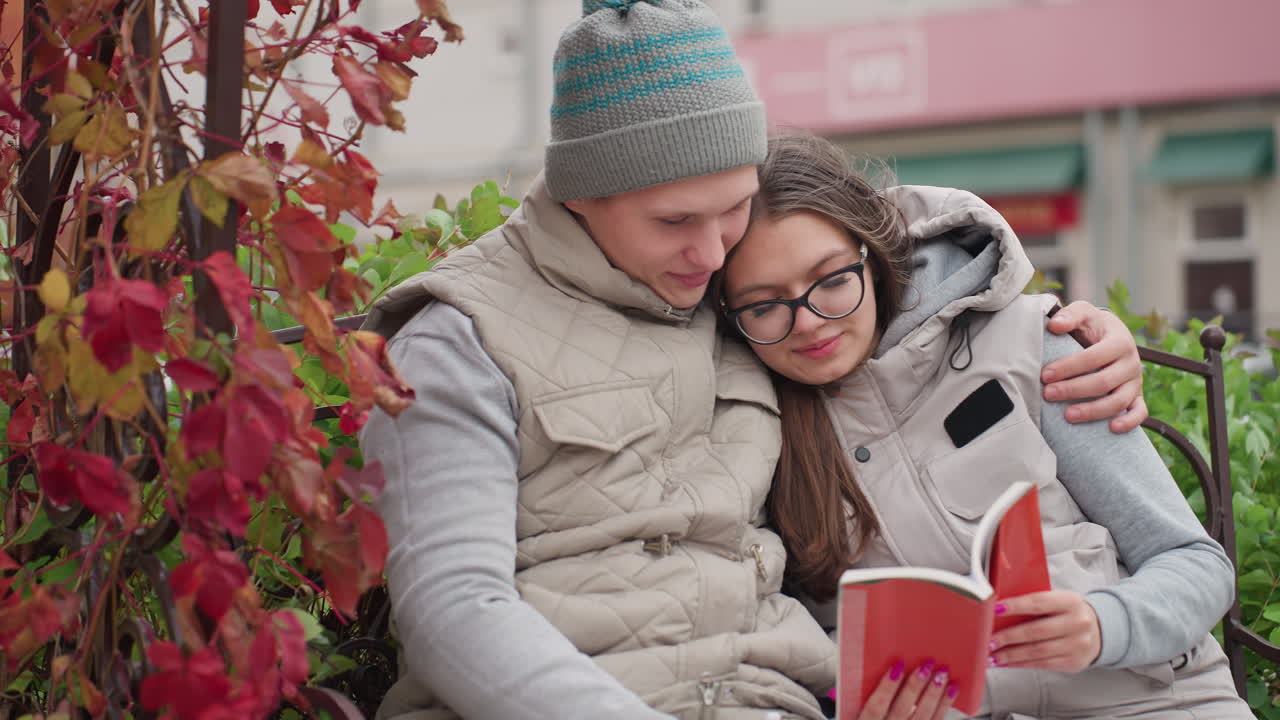 Newly married couple bonding outdoors while sitting on park bench surrounded by red autumn leaves, smiling as wife reads book resting on husband shoulder with blurred background
