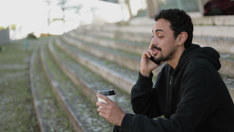 side view of young arabic man with dark curly hair and beard in black hoodie sitting on stairs outside