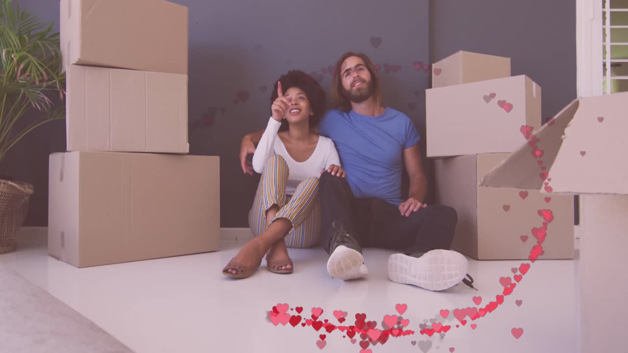 Animación de corazones rojos sobre una pareja feliz y diversa sentada entre cajas después de mudarse a casa