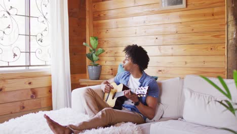 Feliz-Hombre-Afroamericano-Sentado-En-El-Sofá-Y-Tocando-La-Guitarra-En-Una-Cabaña-De-Madera,-Cámara-Lenta