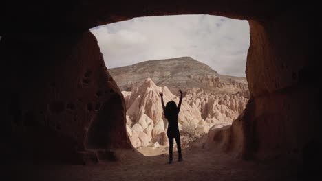 Mujer-Turista-En-Casa-Cueva-Levantando-Los-Brazos-Mientras-Mira-Afuera-En-Formaciones-Rocosas-En-Terreno-árido-De-Capadocia-En-Turquía