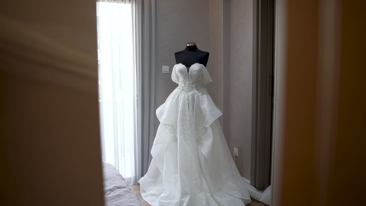 A wedding dress is displayed on a mannequin in a light room. The dress has a flowing design with layers and lace details. Soft light comes through the windows