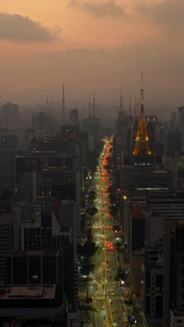 vertical video showing pan right movement over av. paulista, with orange sky, illuminated tower, and vibrant traffic patterns during twilight.