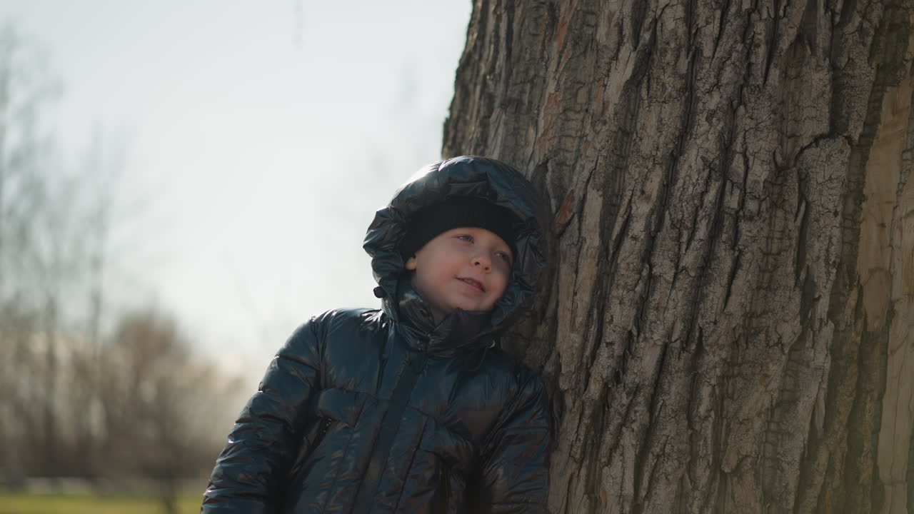 어린 소년은 큰 나무 줄기에 등에 기대어, 부드러운 표정으로 위를 쳐다보고, 그는 반이는 검은색 재을 입고, 배경에는 나무가 있습니다.