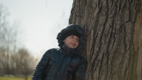 a young boy leaning his back against a large tree trunk, gazing upward with a gentle expression, he is dressed in a shiny black jacket, with trees in the background