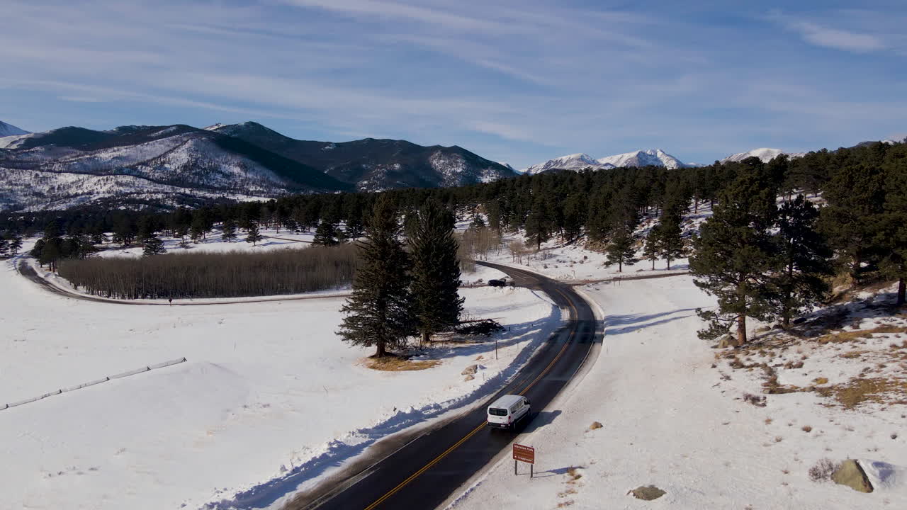 White SUV van drives on snowy mountain pass road through pine tree forest aerial
