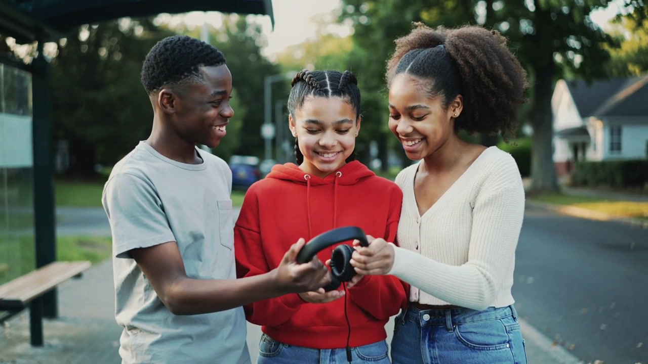 Ein Teenager in einem roten Kapuzenpullover hält ein Telefon und reicht Freunden an einer Bushaltestelle Kopfhörer, um gemeinsam ein Lied zu hören.