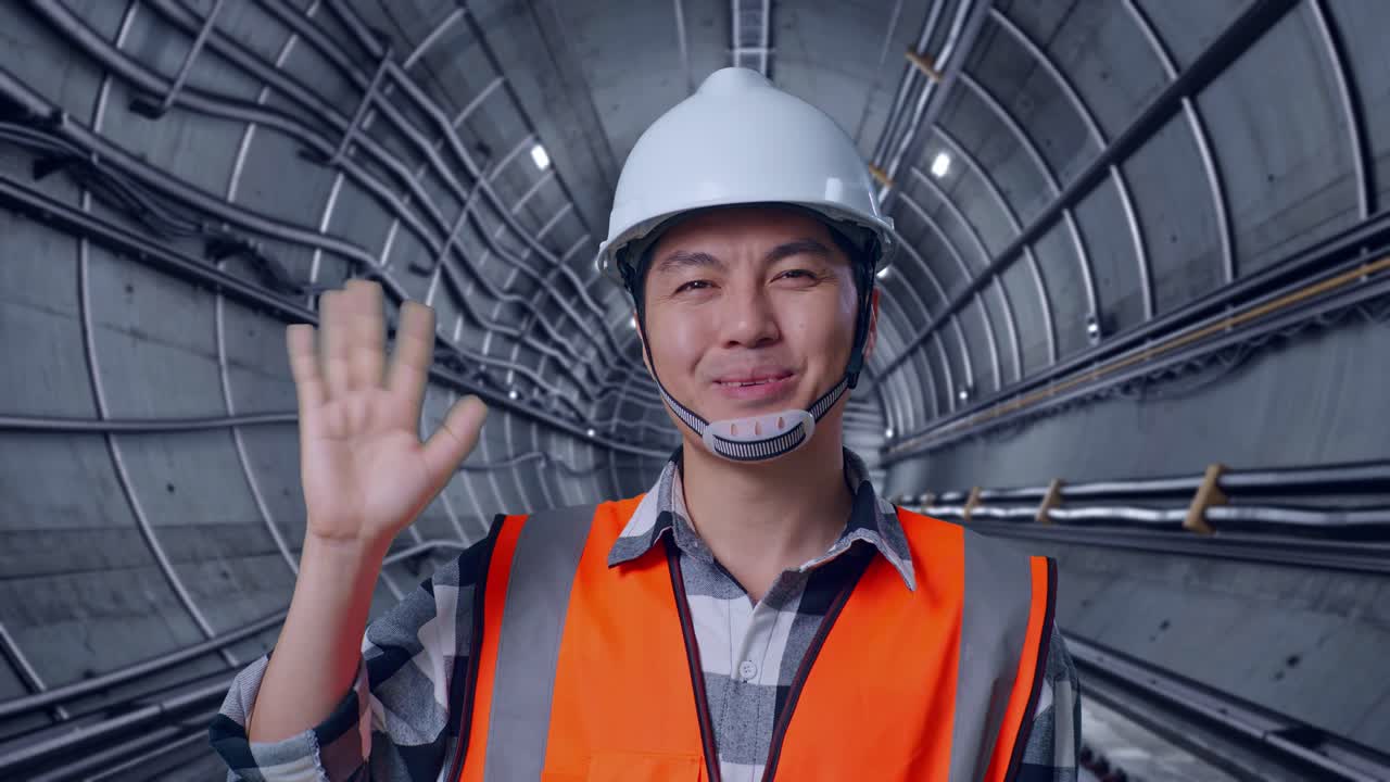 Close Up Of Asian Male Engineer With Safety Helmet Smiling To Camera And Waving Hand Saying Bye While Standing In Underground Subway Tunnel