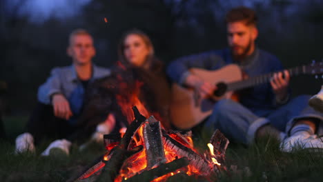 cozy summer evening near campfire in the meadow at forest. young man playing guitar and singing songs and a couple sitting next to him. blurred background.