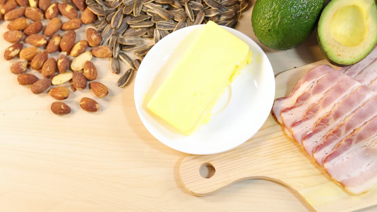 A rotating view of almonds, bacon, butter, avocados, and seeds on a wooden board under bright lighting