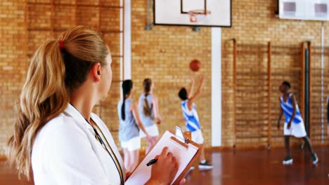 female coach writing on clipboard while students playing in basketball court