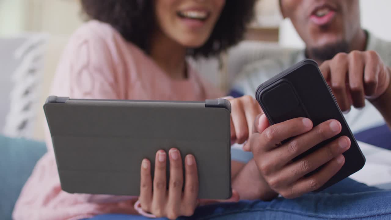 feliz pareja afroamericana usando tableta y teléfono inteligente en la sala de estar