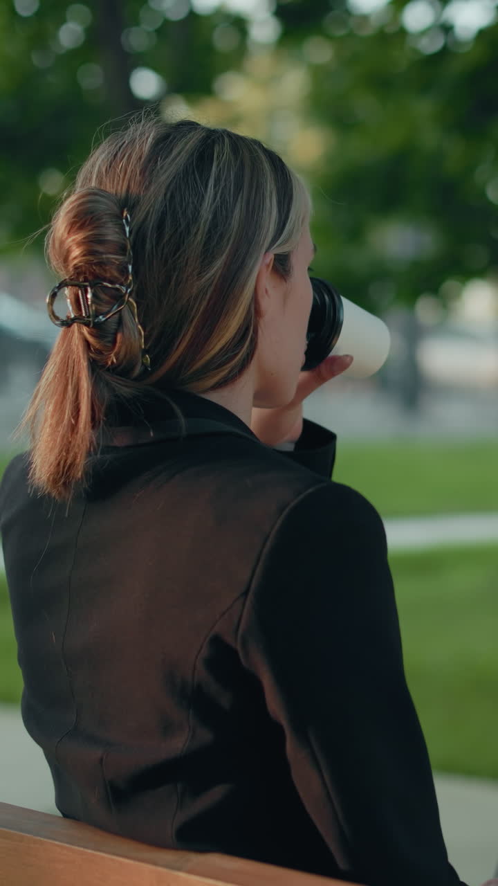 Side view of lady typing on laptop seated on wooden bench in park surrounded by lush greenery with distant parked cars and urban structures visible in blurred background
