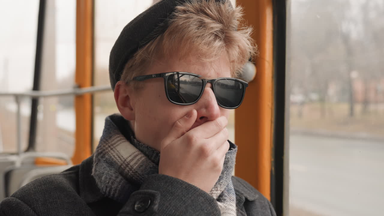 Giovane creativo bianco sul tram, un ritratto contemplativo che guarda attraverso il finestrino macchiato di pioggia. Indossa occhiali da sole, sciarpa di lana e cappotto, mentre la città invernale scorre. Un sottile riflesso sul vetro, in un tragitto lento.