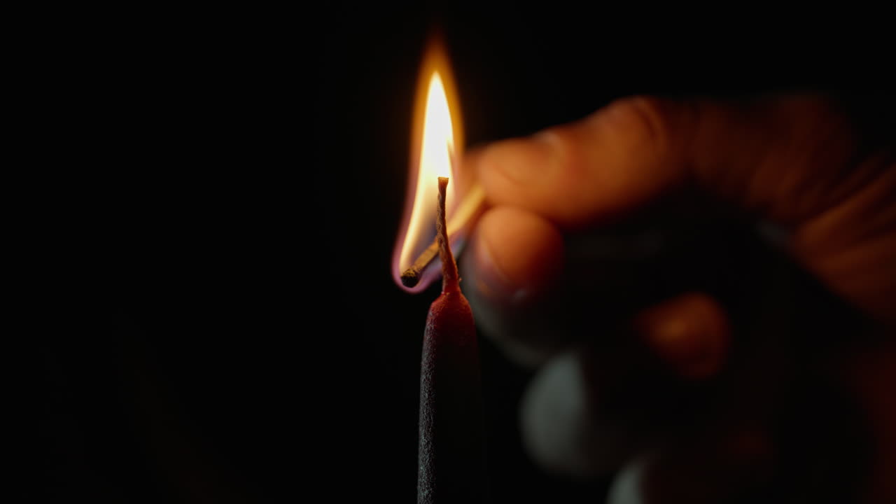 Gros plan d’une main tenant une allumette qui allume une bougie sur une surface noire. Une flamme vive brûle