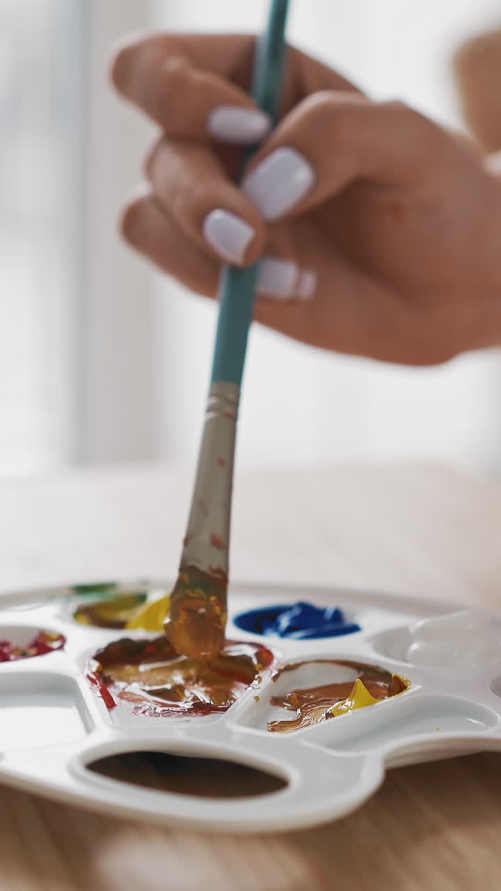 Young woman art student mixes paints to get orange color on white palette with paintbrush on modern wooden easel in studio extreme closeup