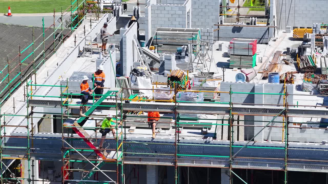 足場やコンクリートブロックの上で作業する高視認性作業着の建設作業員たちの空撮ワイドショット