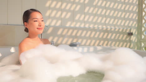 sexy asian woman in jacuzzi bathtub full of foam relaxing with sunlight coming from window, slow motion