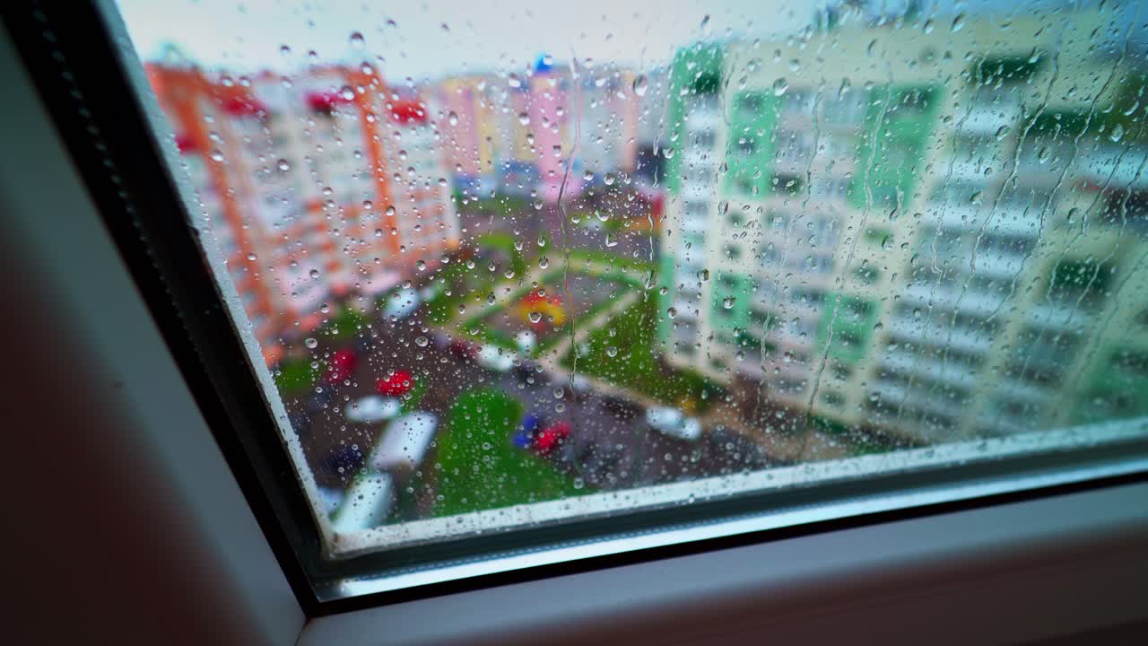 Rainy weather outside. View through the window with raindrops on glass on the background of colorful buildings in the city.