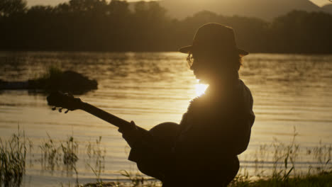 silhouette of musician playing guitar at sunset by the river
