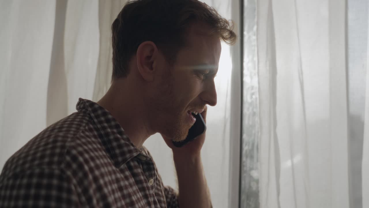 Man making phone call while standing by window, sunlight shining through. Guy talks on his smartphone, frustrated and upset expression, difficult or emotional conversation, stress, conflict, bad news