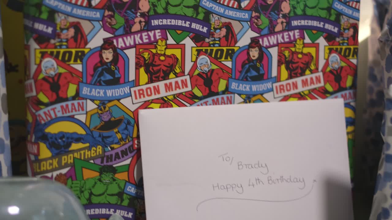 A shot close up and panning across of a young boys birthday presents. This includes presents wrapped in spotty blue paper, football paper and Marvel super heros.